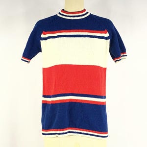 Puede incluir: Una camiseta de punto de manga corta con rayas horizontales azules, blancas y rojas. La camiseta tiene cuello redondo con ribete rojo y blanco, y las mangas también tienen ribete rojo y blanco. La camiseta se muestra en un maniquí.