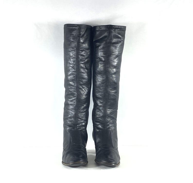 May include: A pair of black knee-high boots featuring a smooth, slightly textured finish. The boots have a tall, structured shaft that reaches the knee, a rounded toe box, and a visible vertical seam running down the center of the front panel.