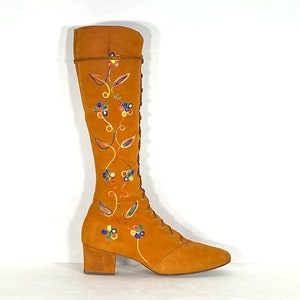 May include: An orange suede knee-high boot with a pointed toe and a block heel. The boot has colourful floral embroidery and a lace-up front. The top of the boot has a decorative trim. The boot is set against a white background.