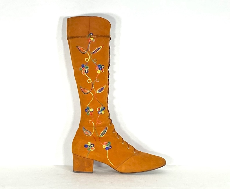 May include: An orange suede knee-high boot with a pointed toe and a block heel. The boot has colourful floral embroidery and a lace-up front. The top of the boot has a decorative trim. The boot is set against a white background.