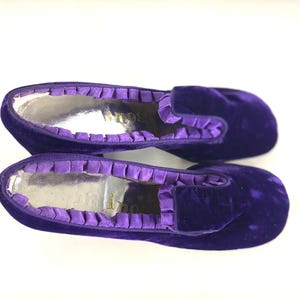May include: A pair of purple velvet slippers with a silver interior. The slippers have a ruffled trim around the opening and a rounded toe. The slippers are a deep shade of purple.