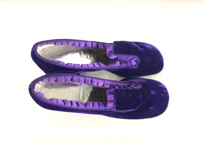 May include: A pair of purple velvet slippers with a silver interior. The slippers have a ruffled trim around the opening and a rounded toe. The slippers are a deep shade of purple.