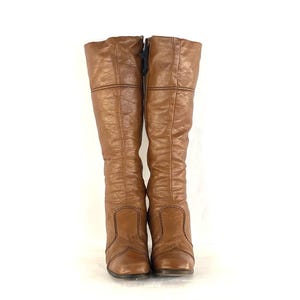 Puede incluir: Un par de botas altas de cuero marr&oacute;n hasta la rodilla. Las botas tienen un ligero tac&oacute;n y un aspecto texturizado. Las costuras negras acent&uacute;an las costuras y la parte superior de la bota. Una cremallera negra es visible en el lateral de una bota.