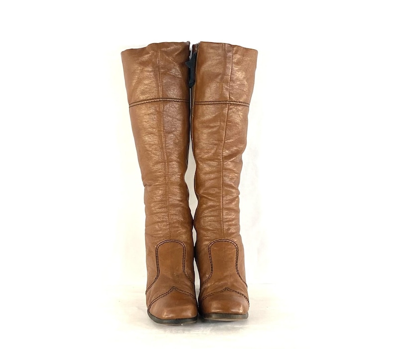 Puede incluir: Un par de botas altas de cuero marr&oacute;n hasta la rodilla. Las botas tienen un ligero tac&oacute;n y un aspecto texturizado. Las costuras negras acent&uacute;an las costuras y la parte superior de la bota. Una cremallera negra es visible en el lateral de una bota.