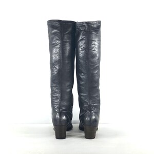 May include: A pair of knee-high black boots with a textured finish and a smooth, rounded toe. The boots feature a vertical seam running down the center of the shaft and a sturdy, block-style heel. The material has a slight sheen and visible creases.