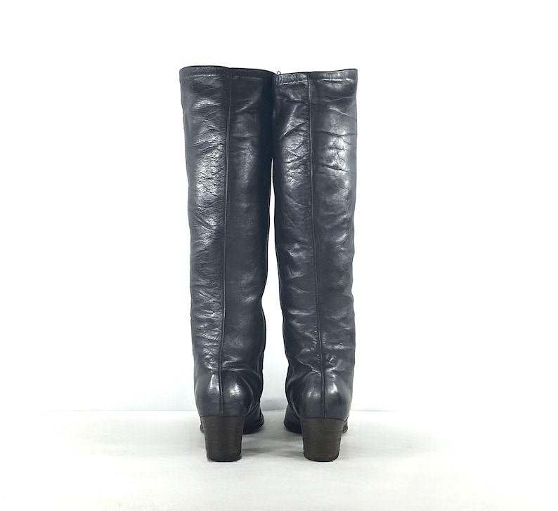 May include: A pair of knee-high black boots with a textured finish and a smooth, rounded toe. The boots feature a vertical seam running down the center of the shaft and a sturdy, block-style heel. The material has a slight sheen and visible creases.
