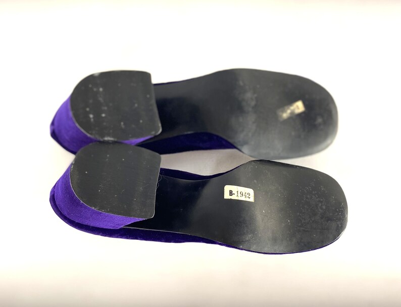 May include: A pair of purple velvet shoes with black soles. The shoes have a low heel and a rounded toe. The soles have a sticker with the number B-1942.