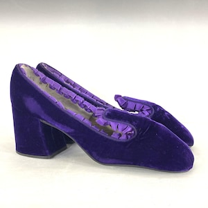 May include: A pair of vintage purple velvet heeled shoes. The shoes have a square toe and a block heel. The top of the shoe has a ruffled trim. The shoes are approximately 3 inches high.