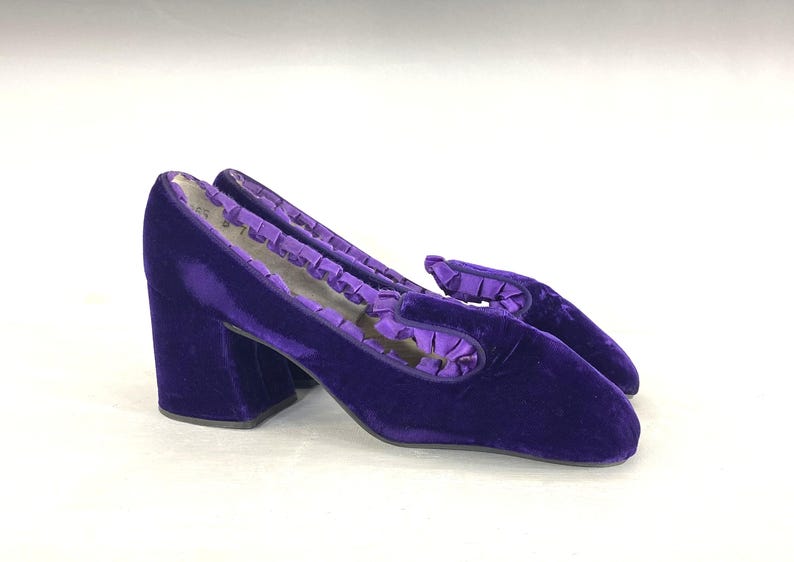 May include: A pair of vintage purple velvet heeled shoes. The shoes have a square toe and a block heel. The top of the shoe has a ruffled trim. The shoes are approximately 3 inches high.