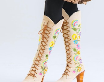 Puede incluir: Botas beige hasta la rodilla con cordones marrones en la parte delantera, tacón de madera apilada y bordados florales vibrantes en tonos azul, amarillo, rosa y verde en los laterales. Las botas tienen punta afilada y una caña alta.