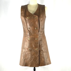 May include: A sleeveless brown leather dress featuring a double-breasted front closure with six small gold-toned metal buttons. The garment has a V-neckline, a fitted bodice, and a slightly flared skirt, showcasing a smooth, polished texture.