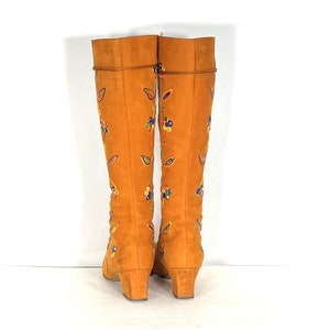 May include: A pair of knee-high orange suede boots with embroidered floral and leaf designs in various colours. The boots have a small heel and a rounded toe. The boots are displayed against a white background.
