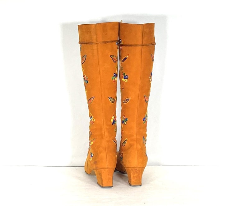 May include: A pair of knee-high orange suede boots with embroidered floral and leaf designs in various colours. The boots have a small heel and a rounded toe. The boots are displayed against a white background.