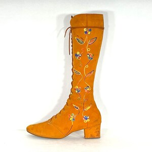 May include: An orange suede knee-high boot with a lace-up front and a small block heel. The boot features embroidered floral and leaf designs in blue, yellow, and green. The boot's shaft is approximately 38 cm high.