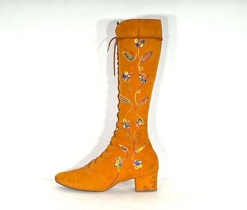 May include: An orange suede knee-high boot with a lace-up front and a small block heel. The boot features embroidered floral and leaf designs in blue, yellow, and green. The boot's shaft is approximately 38 cm high.
