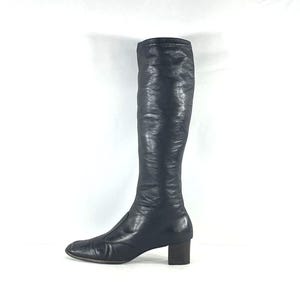 May include: A knee-high black leather boot with a smooth, slightly textured finish, a square toe, and a low, block-shaped heel. The boot features a clean, minimalist silhouette with a vertical seam running down the front of the shaft.
