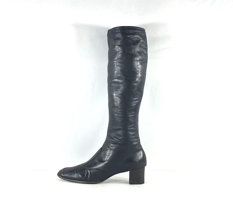 May include: A knee-high black leather boot with a smooth, slightly textured finish, a square toe, and a low, block-shaped heel. The boot features a clean, minimalist silhouette with a vertical seam running down the front of the shaft.