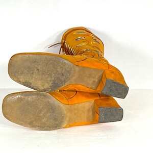 May include: Mustard-yellow suede boots with a square toe and block heel. The boots have decorative stitching and fringe detailing. The soles show wear. The boots are approximately 30 cm high.