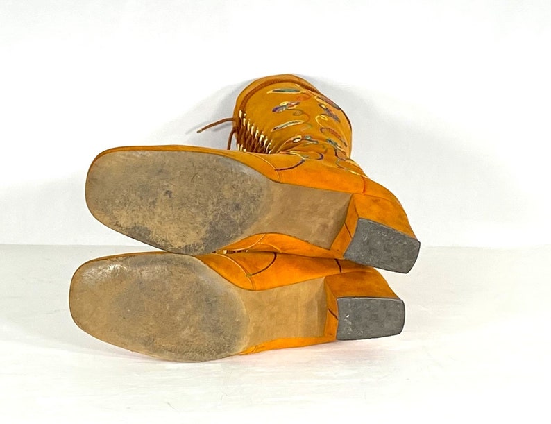 May include: Mustard-yellow suede boots with a square toe and block heel. The boots have decorative stitching and fringe detailing. The soles show wear. The boots are approximately 30 cm high.