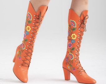 Puede incluir: Un par de botas naranjas de caña alta con bordados florales y cordones en la parte delantera. Las botas tienen un tacón grueso.