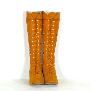 May include: A pair of knee-high, orange suede boots with a lace-up design. The boots feature decorative floral and leaf patterns in various colours. The top edge has a scalloped design. The boots are displayed against a white background.