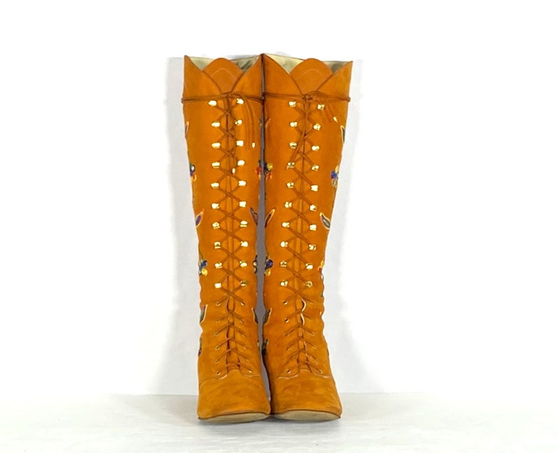 May include: A pair of knee-high, orange suede boots with a lace-up design. The boots feature decorative floral and leaf patterns in various colours. The top edge has a scalloped design. The boots are displayed against a white background.
