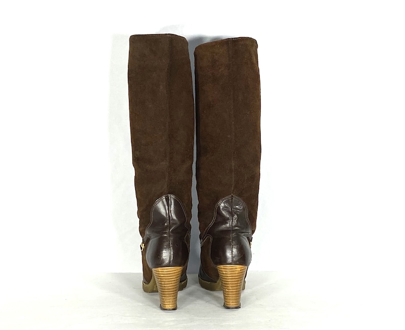 May include: A pair of brown suede knee-high boots with a wooden heel. The boots have a zipper on the inside of the leg and a buckle on the outside.