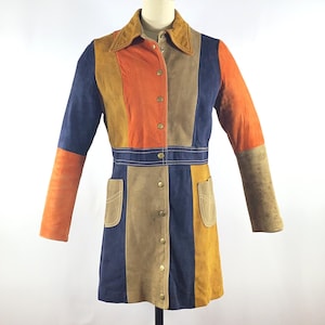 May include: A long-sleeved jacket featuring a color-blocked design in shades of navy blue, burnt orange, and tan. The garment has a pointed collar, a front button closure with metallic buttons, and two patch pockets on the lower front section.