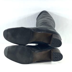 May include: A pair of black leather knee-high boots with square toes and block heels. The outsoles are black with a fine horizontal ribbed texture, while the heels feature a dark brown stacked appearance with a textured, grid-patterned rubber base.
