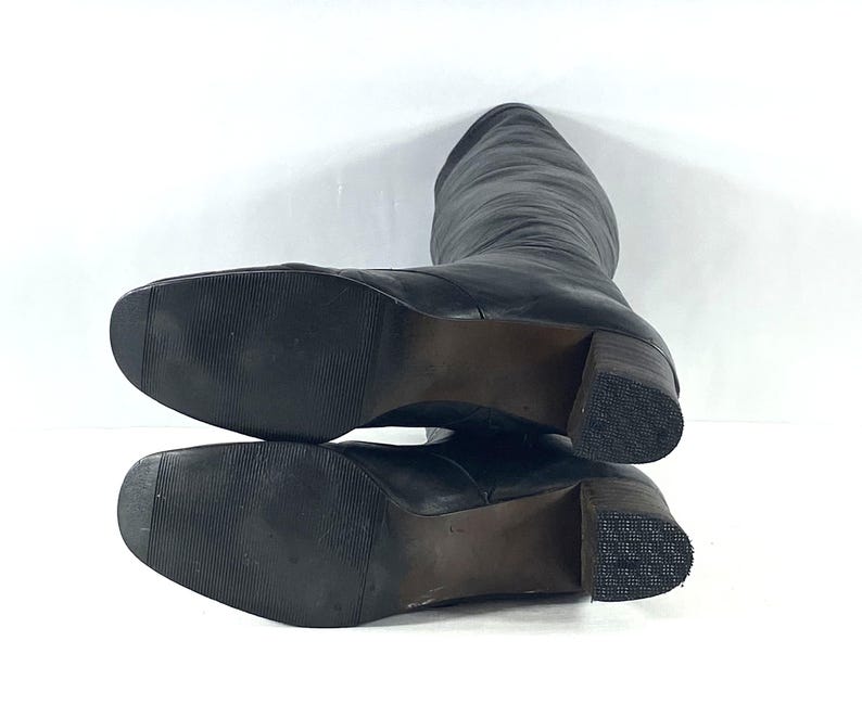 May include: A pair of black leather knee-high boots with square toes and block heels. The outsoles are black with a fine horizontal ribbed texture, while the heels feature a dark brown stacked appearance with a textured, grid-patterned rubber base.