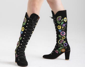 Puede incluir: Botas negras de caña alta con un diseño de bordado floral. Las botas tienen un cierre de encaje en la parte delantera y un tacón grueso.