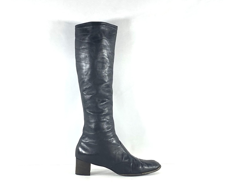 May include: A knee-high black leather boot with a smooth, slightly textured finish and a rounded toe. The boot features a block heel and a streamlined silhouette that extends from the foot up to the calf, showcasing a consistent dark tone.