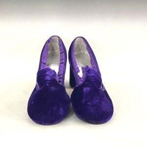 May include: A pair of vintage purple velvet heels. The shoes have a rounded toe, a thick heel, and a ruffled trim around the opening. The velvet material gives the shoes a luxurious look. The heels are a classic style.