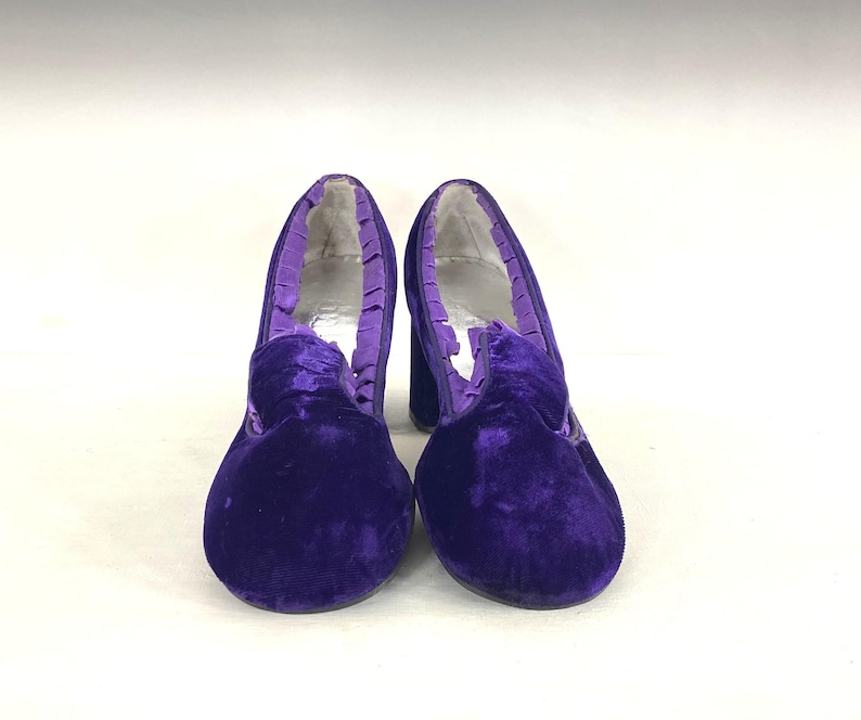 May include: A pair of vintage purple velvet heels. The shoes have a rounded toe, a thick heel, and a ruffled trim around the opening. The velvet material gives the shoes a luxurious look. The heels are a classic style.