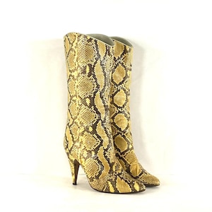 Pode incluir: Um par de botas altas com salto e padrão de pele de cobra em tons de amarelo, castanho e preto. As botas têm bico fino e salto agulha. As botas têm cerca de 30 cm de altura.