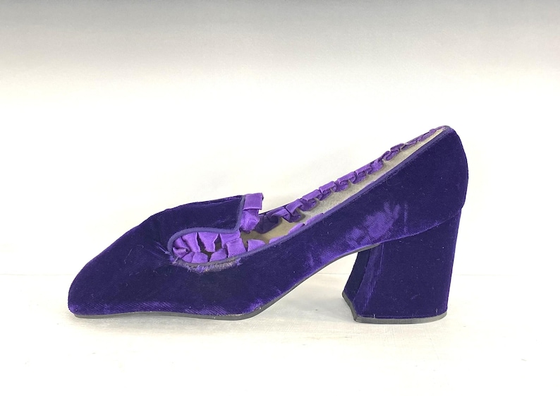 May include: A single, vintage, purple velvet shoe with a block heel. The shoe has a rounded toe and a decorative ruffle detail on the vamp. The heel measures approximately 3 inches.