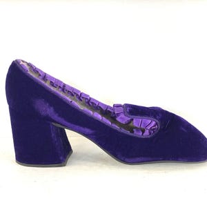 May include: A single, vibrant purple velvet shoe with a block heel. The shoe features a rounded toe and a ruffled trim along the opening. The heel is approximately 3 inches high. The shoe is set against a white background.