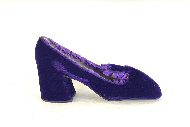 May include: A single, vibrant purple velvet shoe with a block heel. The shoe features a rounded toe and a ruffled trim along the opening. The heel is approximately 3 inches high. The shoe is set against a white background.