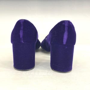 May include: A pair of purple velvet block-heeled shoes. The shoes have a rounded toe and a thick heel. The velvet material gives the shoes a luxurious look. The heels are approximately 3 inches high.