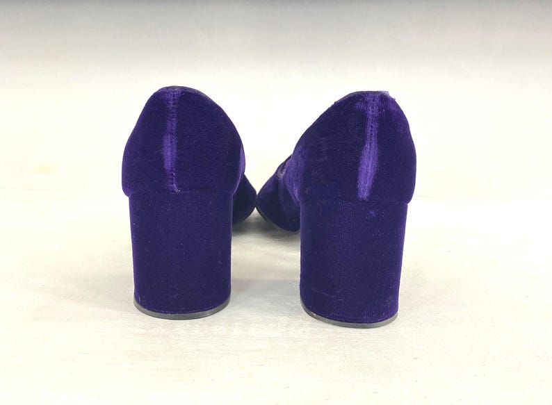 May include: A pair of purple velvet block-heeled shoes. The shoes have a rounded toe and a thick heel. The velvet material gives the shoes a luxurious look. The heels are approximately 3 inches high.