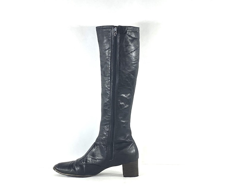 May include: A knee-high black leather boot with a smooth, slightly textured finish. The boot features a full-length vertical zipper along the side, a rounded toe, and a sturdy, block-style heel. The design is sleek, simple, and functional.