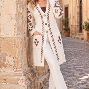 May include: A long-sleeved, cream-colored knit cardigan featuring a V-neckline, three silver-toned buttons, and two front pockets. The garment is decorated with brown geometric triangle patterns along the sleeves, pockets, and front placket.