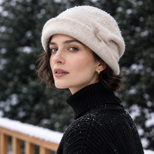May include: A beige felted wool cloche hat with a rolled brim and a small gold-toned metal chain accent on the side. The hat features a textured surface and is worn with a black ribbed turtleneck sweater.