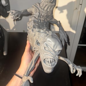May include: A gray 3D printed figurine of a creature with a long, segmented body, sharp teeth, and multiple limbs. The figure features a detailed, textured surface with a prominent head, wide mouth, and clawed hands extending outward.
