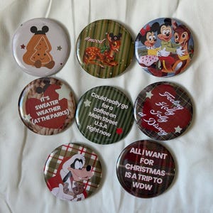 May include: Eight circular pinback buttons featuring Disney-themed holiday designs. Graphics include a gingerbread cookie, Bambi, Mickey and Minnie Mouse, Goofy, and text such as 'It's sweater weather', 'Feeling holly jolly', and 'All I want for Christmas is a trip to WDW'.