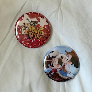 May include: Two circular pin-back buttons. One button features a red bandana pattern with a white star and the text Frontierland. The second button depicts two cartoon mice wearing cowboy hats and western attire against a light blue background.
