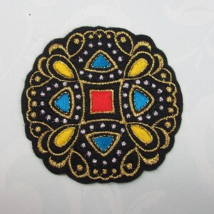 May include: A black felt patch with a gold and silver embroidered design. The design features a central square with a red square in the center, surrounded by four blue triangles. The design is surrounded by a border of gold and silver swirls.
