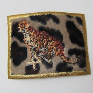 May include: Embroidered cheetah patch with a gold border. The cheetah is running and has a brown, orange, and white color scheme. The background is a brown and tan leopard print.