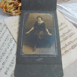 May include: A sepia-toned vintage photograph of a woman in a dark dress, presented in a dark grey photo album. The album is open, showing the portrait. Musical notation and a decorative flower are in the background.