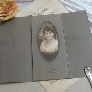 May include: A sepia-toned photograph of a woman in a white dress and pearl necklace, set in an oval frame within a gray, tri-fold photo holder. The holder has embossed floral designs. A vintage rose and old papers are in the background.